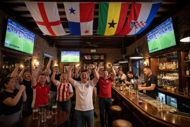 People in a bar cheering with the flags of Englands World Cup 2026 group and sports broadcasts on screens.